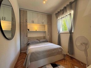 A bedroom with a bed, a mirror, a window with curtains, a fan, and built-in cabinets. - Holiday apartment mit Meerblick (Dramalj)