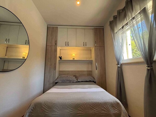 Une chambre à coucher avec un lit, un miroir, une commode, une étagère avec une lampe et une fenêtre avec des rideaux.
