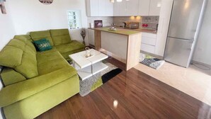Un espace de vie compact avec un canapé vert, une petite table basse, un coin cuisine doté de meubles blancs, un réfrigérateur et des luminaires suspendus.