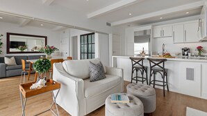 A modern living room with a sofa, ottoman, and bar stools. There is a dining area with a table and chairs. The kitchen is visible with white cabinets and a countertop.
