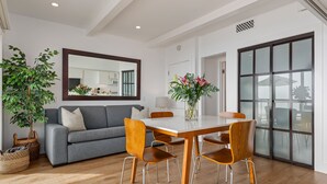 A modern living room with a grey sofa, wooden dining table, and chairs. There is a large mirror on the wall, a potted plant, and a window with a view.
