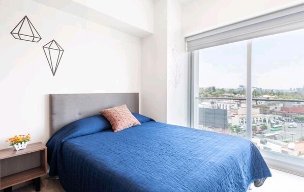 A bedroom with a large window, a bed with blue bedding, a nightstand with a vase of flowers, and geometric wall art.