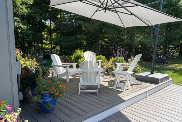 An outdoor deck with white chairs and a large umbrella, surrounded by potted plants and a wooden deck.