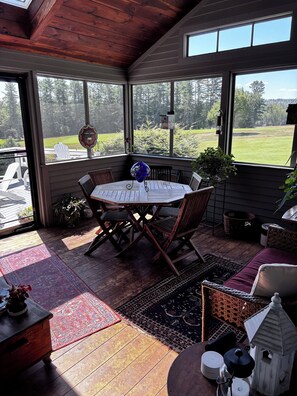 A covered patio with a wooden table and chairs, surrounded by large windows offering a view of a green landscape.