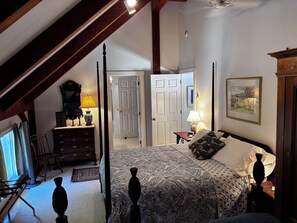 A bedroom with a large bed, a dresser, a chair, and a desk with a lamp. There is a door leading to another room.