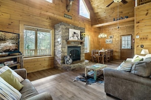 A cozy log cabin interior with a stone fireplace, wooden furniture, and a dining area.