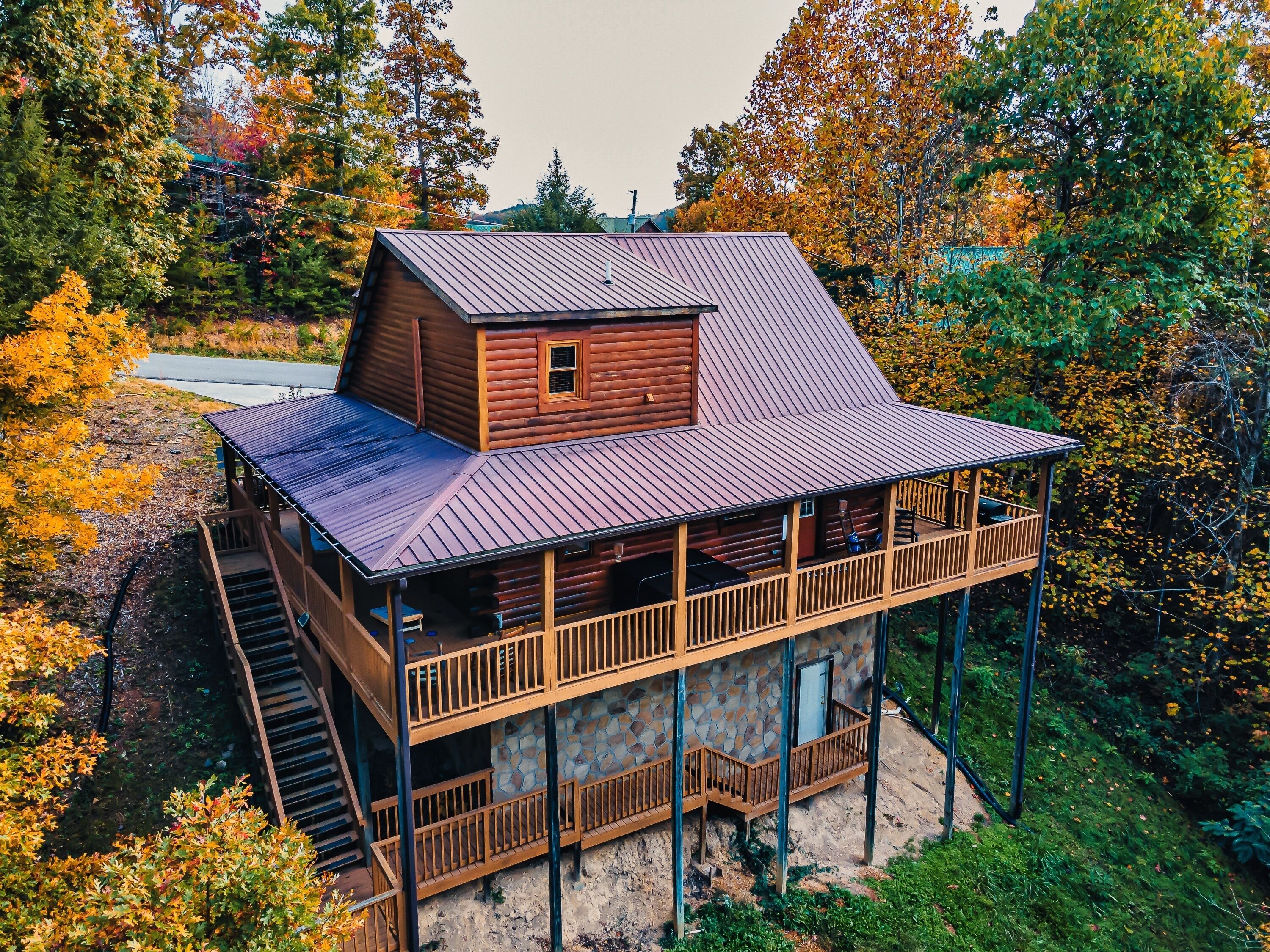 A two-story log cabin with a balcony and a small attached shed, surrounded by autumn foliage.