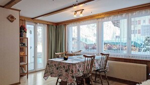 Comedor con una mesa dispuesta para cuatro personas, sillas de madera, una estantería y ventanas grandes con cortinas.