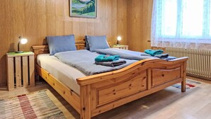 Una cama de madera con cabecera, dos mesitas de noche con lámparas, un cuadro enmarco con un paisaje, y una ventana con cortinas de gasa.