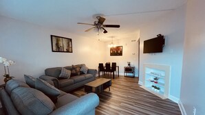 Ein Wohnzimmer mit grauem Sofa, einem Couchtisch, einem Essbereich mit Stühlen, einem Deckenventilator, einem Kamin und einem Wand-Fernseher.