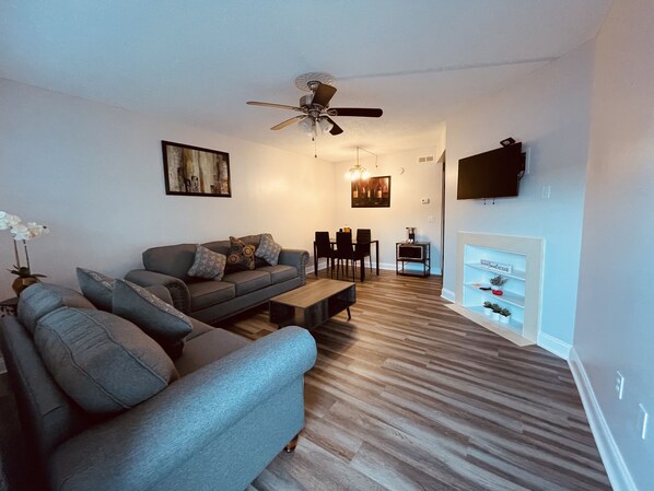 A living room with a grey sofa, a coffee table, a dining area with chairs, a ceiling fan, a fireplace, and a mounted TV.