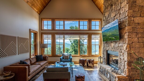 A spacious living room with a high ceiling, large windows, a stone fireplace, and a mounted television.