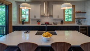 A modern kitchen with a large island, two pendant lights, and a view of greenery outside.