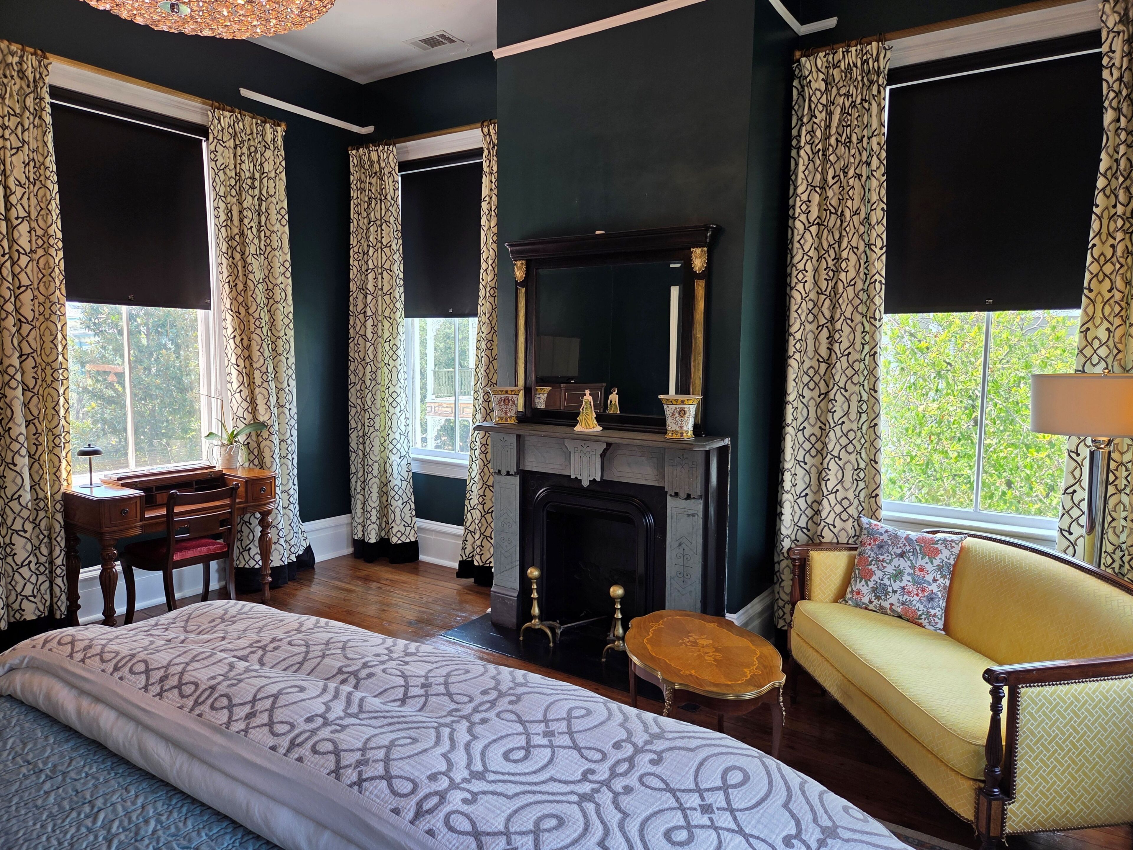 A room with a bed, a sofa, a fireplace, a desk, and large windows with patterned curtains.