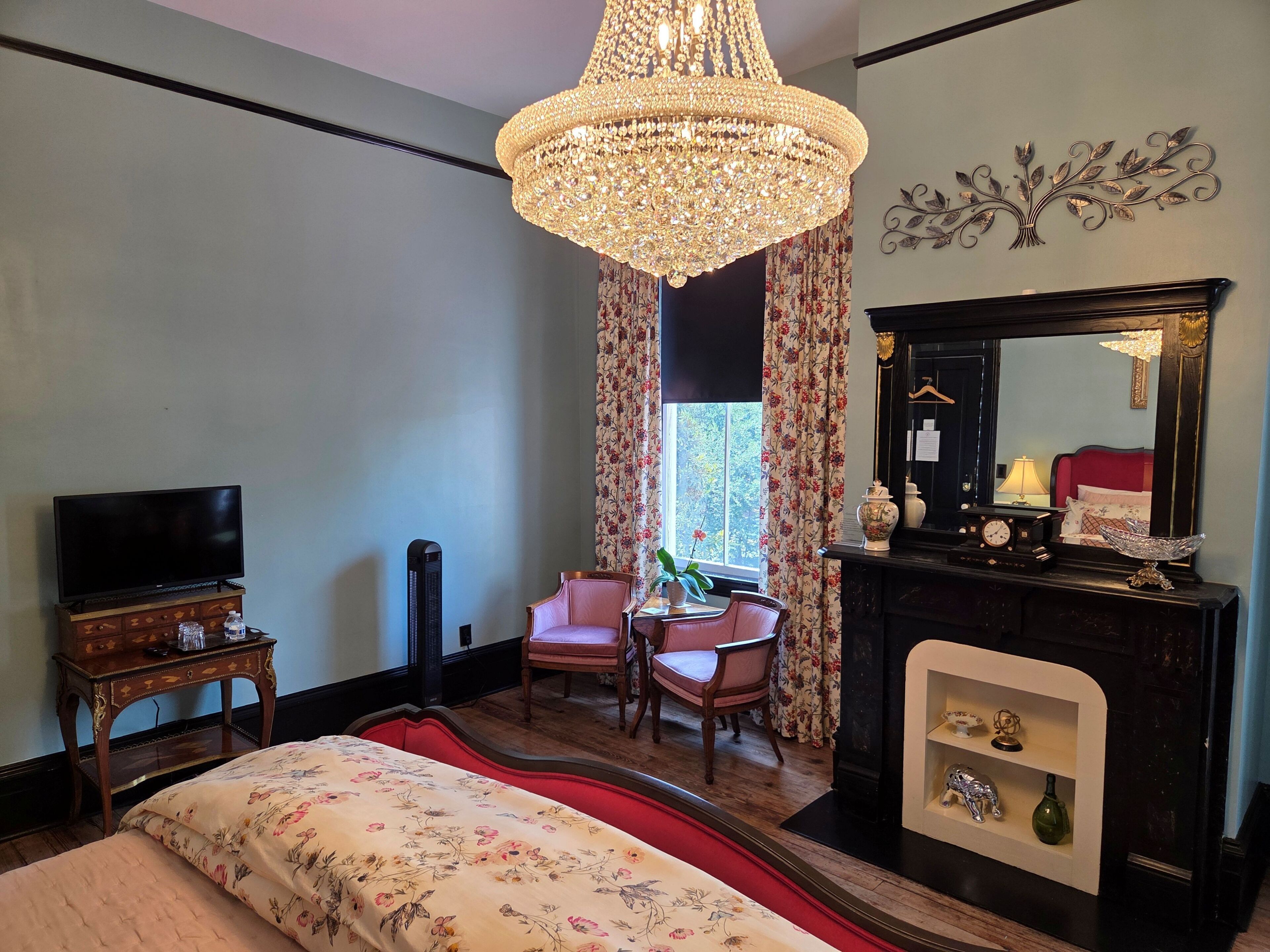 A bedroom with a bed, a television, a desk, a chair, a fireplace, and a chandelier.