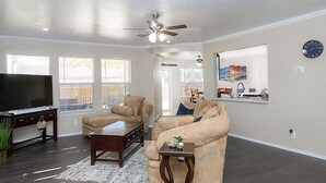 A spacious living room with a large flat-screen TV, two beige sofas, a coffee table, and a ceiling fan.