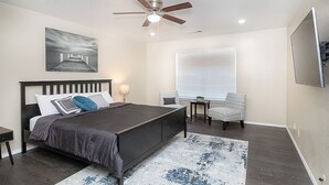 A bedroom with a bed, a ceiling fan, a window with blinds, a chair, a small table, and a painting on the wall.