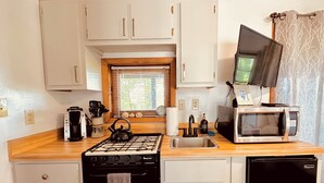 Eine kompakte Küche mit Gasherd, Mikrowelle, Spüle und kleinem Kühlschrank. Ein Fenster mit Jalousien, ein Wasserkocher und eine Kaffeemaschine befinden sich auf der Arbeitsfläche.