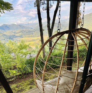Un fauteuil suspendu en osier, doté d’un coussin, offrant une vue sur un paysage montagneux verdoyant et un ciel dégagé.