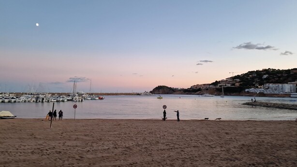 A beach with a marina, boats, and a few people walking.