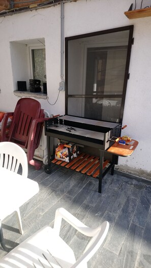 Um pátio externo com uma churrasqueira, uma cadeira branca, uma cadeira vermelha e uma janela com vista.