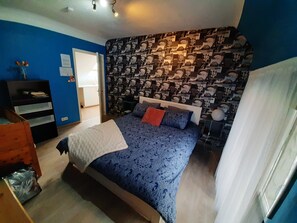 Una camera da letto con parete decorata, un letto con lenzuola blu, una cassettiera in legno e una porta che dà su un'altra stanza.