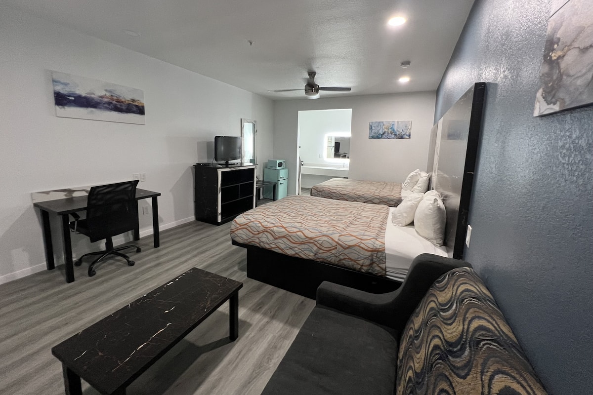 A hotel room with a bed, a desk, a chair, a bench, a ceiling fan, and artwork on the walls.