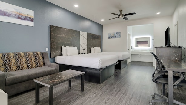 A modern hotel room with a grey sofa, a bed with white bedding, a small table, a desk with a chair, and a ceiling fan.