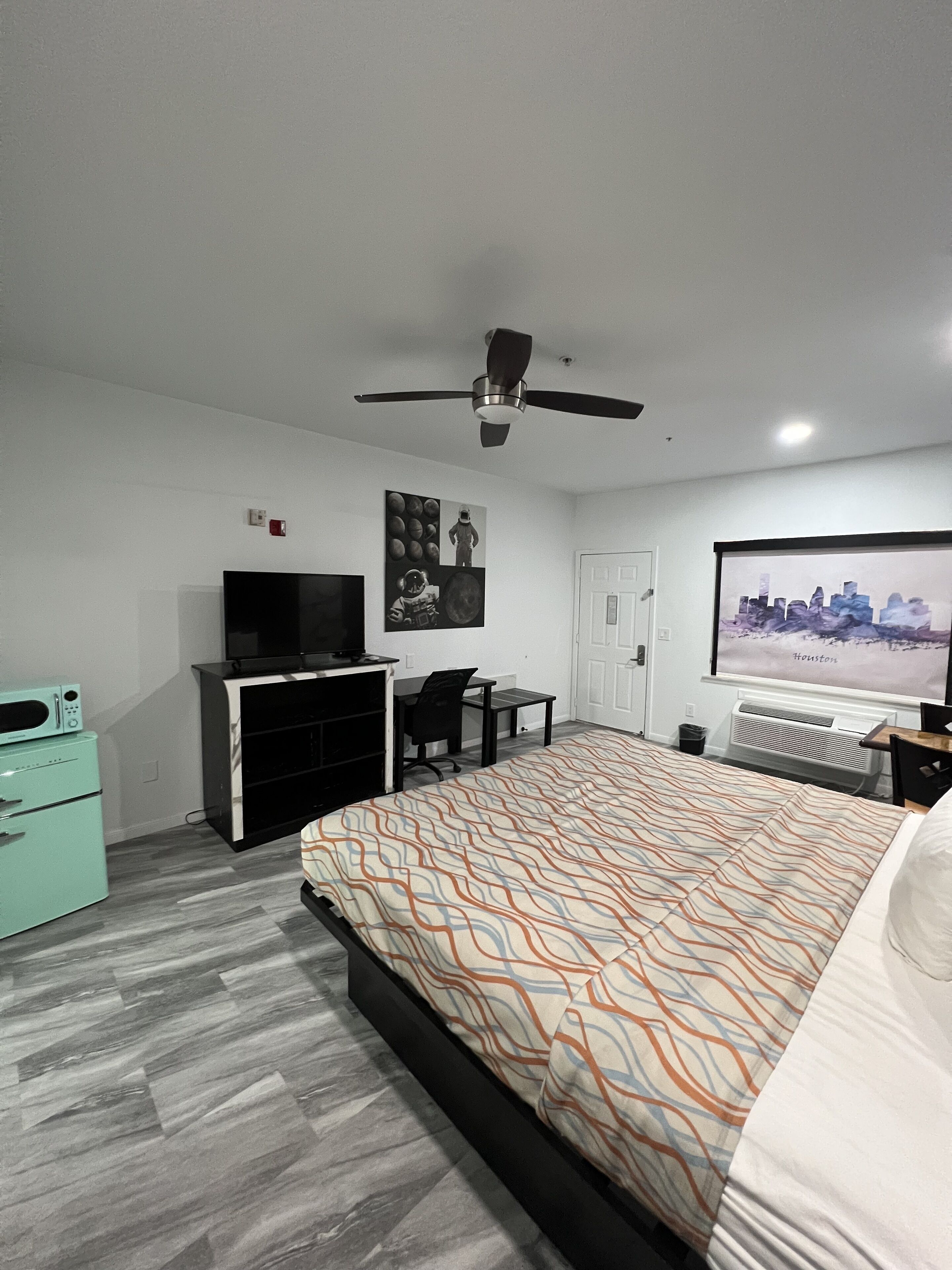 A hotel room with a bed, a desk, a television, a ceiling fan, and a cityscape poster on the wall.