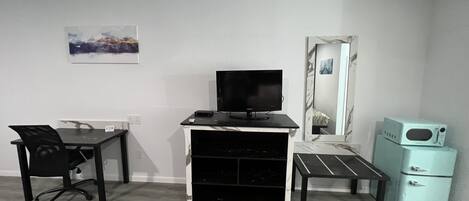 A room with a desk, chair, TV, microwave, and a mirror.