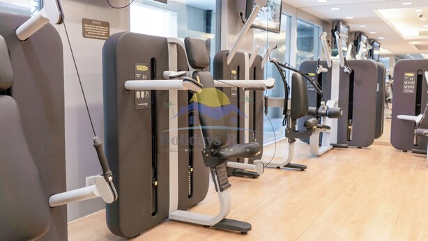 A row of exercise machines in a gym with a wooden floor and a glass wall in the background. - Holidays R Us villa 3 bedroom premium location (Dubai)