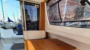 A boat interior with a wooden table and cushioned seating, a window with a view of buildings, and a ventilation system. - UNUSUAL NIGHT ABOARD A BOAT (Saint-Florent)