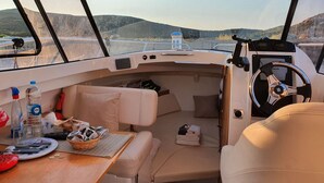 A boat interior with a dining area, cushioned seating, a steering wheel, and a view of the water and hills. - UNUSUAL NIGHT ABOARD A BOAT (Saint-Florent)