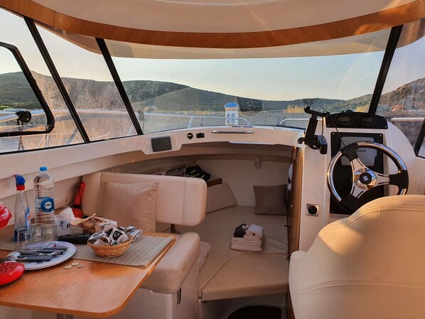 A boat interior with a dining area, cushioned seating, a steering wheel, and a view of the water and hills.