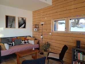 A cozy living room with a wooden wall, a sofa with patterned cushions, a bench, a chair, a bookshelf, and a window with a view of a snowy landscape.