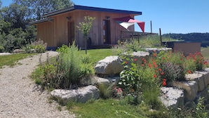 Eine Holzhütte mit einer Veranda, umgeben von einem Garten mit verschiedenen Pflanzen und Blumen.