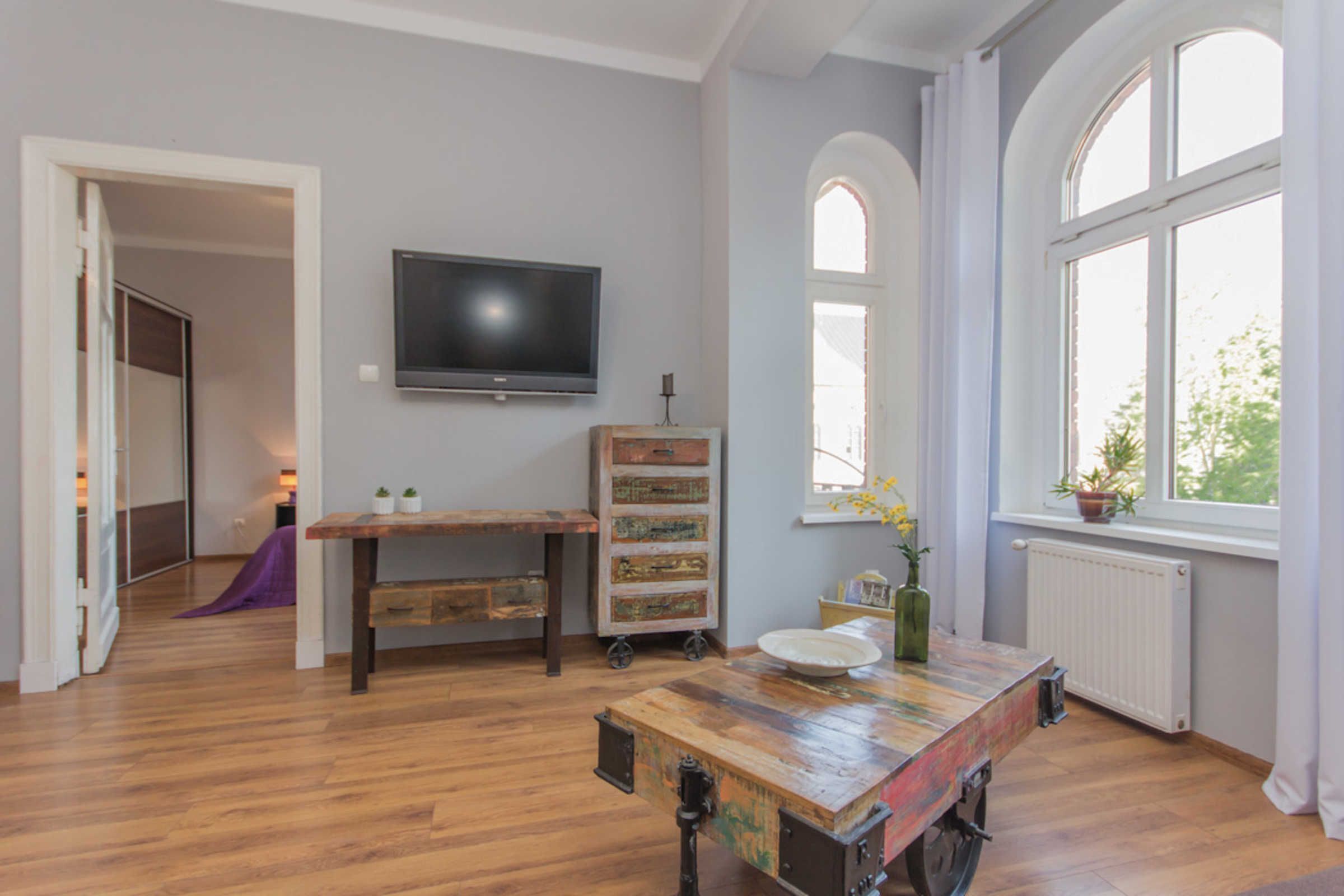Ein Zimmer mit Holzboden, einem an der Wand montierten Fernseher, einem Fenster mit Blick auf Grünflächen und einem Tisch mit einer Vase.