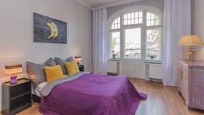 Un dormitorio con una cama grande, una ventana con cortinas, una mesita de noche con lámpara y una obra de arte en la pared.