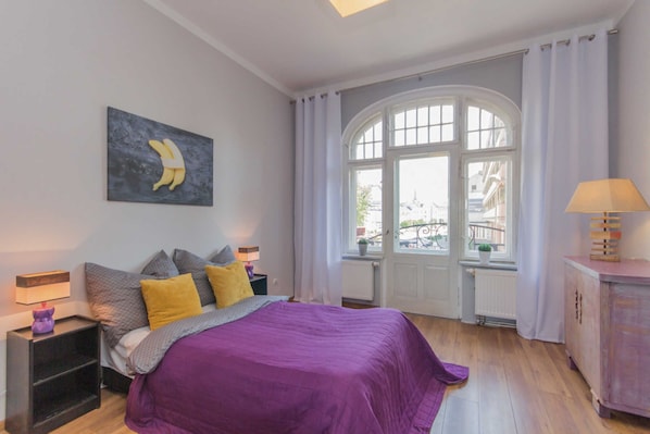 Une chambre à coucher avec un grand lit, une fenêtre avec des rideaux, une table de chevet avec une lampe et une œuvre d’art accrochée au mur.