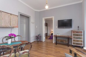 Un salon avec un plancher en bois, une table à manger avec des chaises, un téléviseur fixé au mur et une petite table sur laquelle est posée une plante.