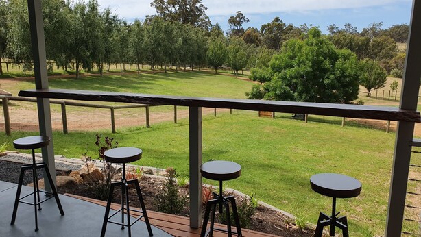 A covered outdoor seating area with three stools and a table, overlooking a grassy landscape with trees and a fence.