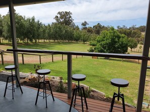 A covered outdoor seating area with three stools and a table, overlooking a grassy landscape with trees and a fence.