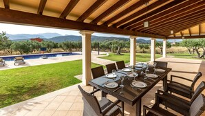 An outdoor patio with a dining table set for six, surrounded by wicker chairs, under a wooden pergola with a view of a swimming pool and mountains in the background. - Modern finca, secluded location, 10min to the dream beach Playa del Muro, large pool (Búger)