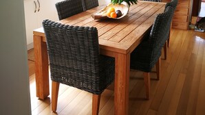 Uma mesa de jantar de madeira com cadeiras de vime, um vaso de flores e uma fruteira.