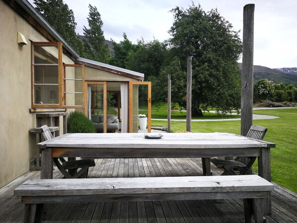 A wooden deck with a table and benches, a glass door leading to a room, and a view of a grassy area with trees and hills in the background.