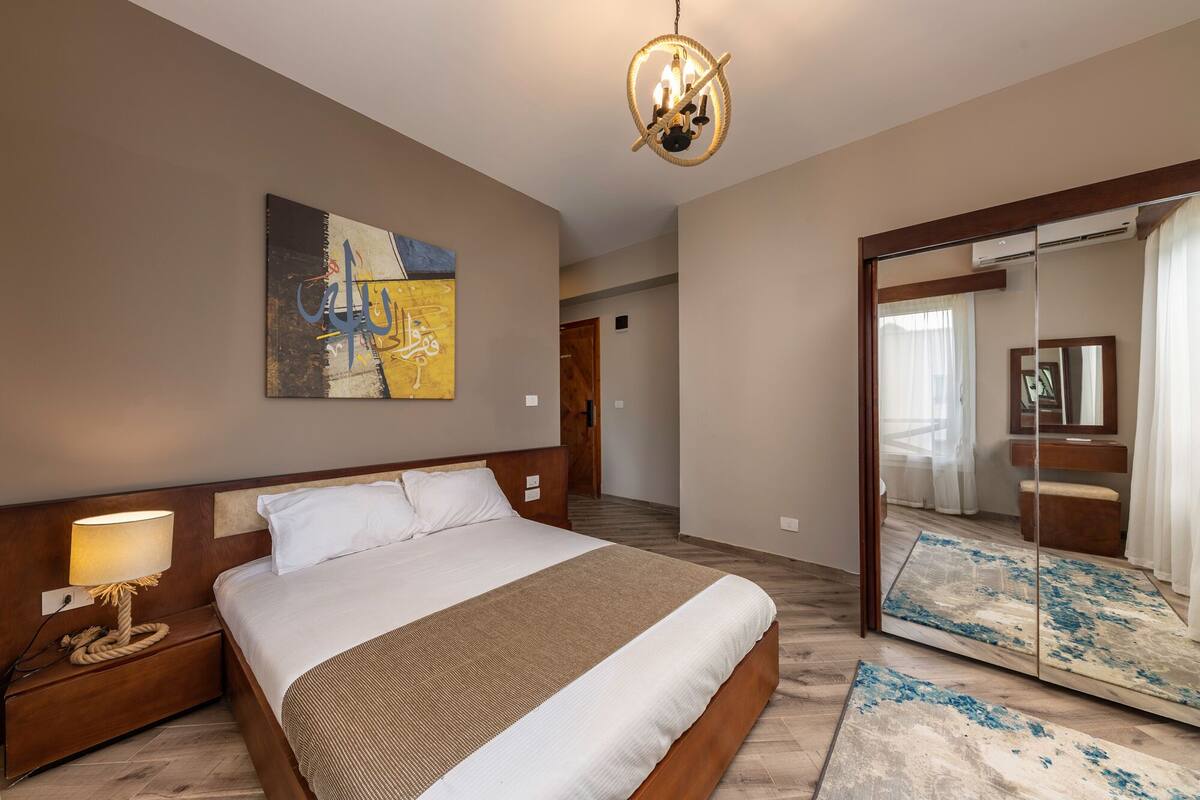 A hotel room with a large bed, a bedside lamp, a painting on the wall, and a mirror with a rug.