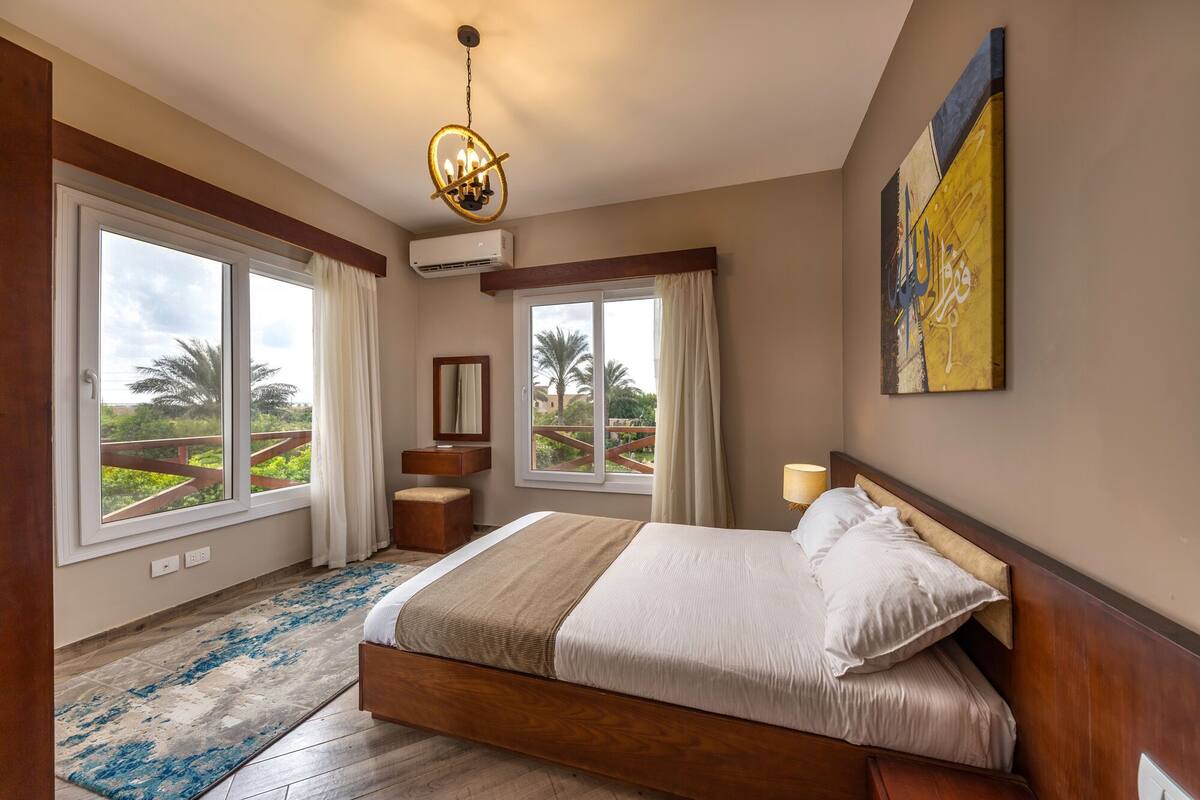 A bedroom with a large bed, a nightstand, a mirror, and a view of palm trees through large windows.