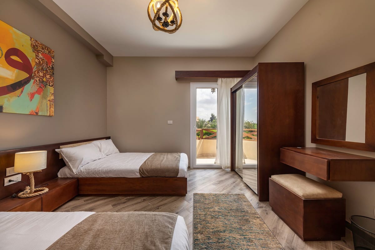 A hotel room with two beds, a large window, a wooden desk, and a ceiling light.