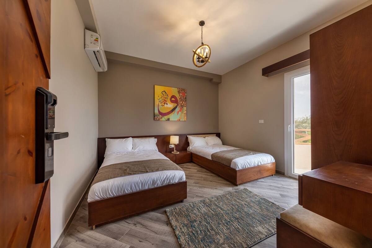 A bedroom with two beds, a ceiling light fixture, a painting on the wall, and a sliding door leading to an outdoor area.