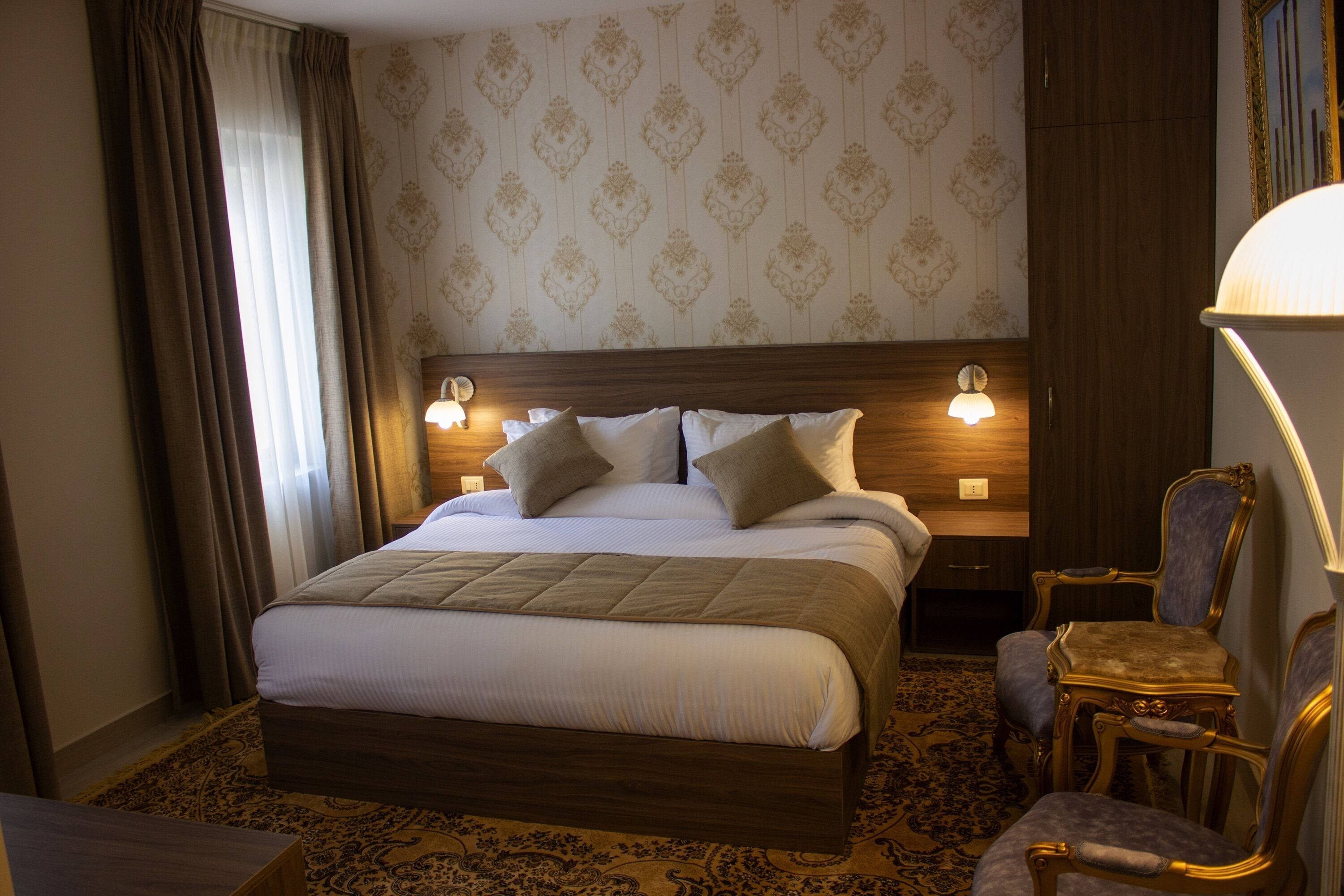 Habitación de hotel con una cama grande, dos lámparas de noche, cabecera de madera, alfombra estampada y una silla con lámpara.
